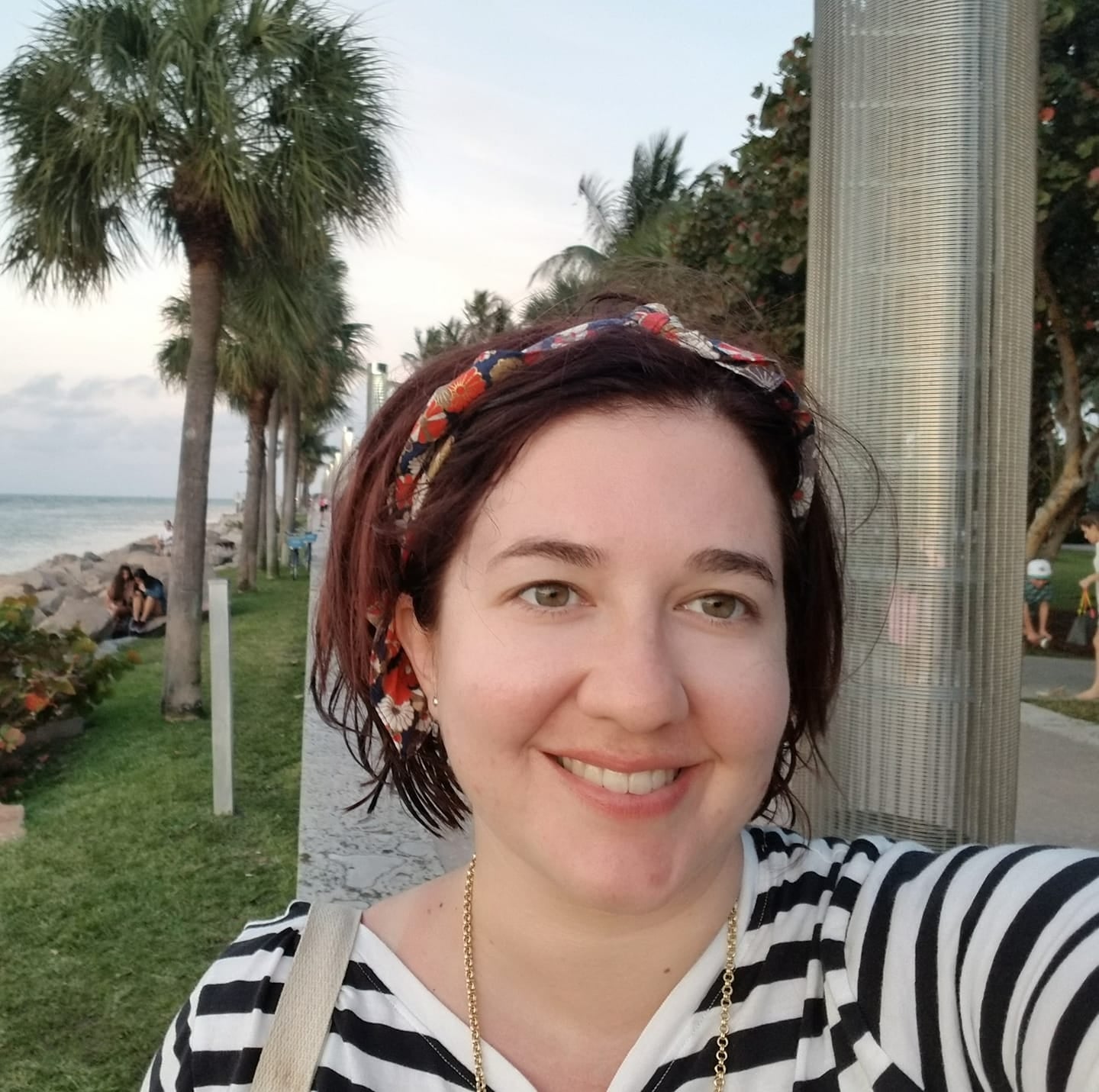 A smiling woman, Bethany, in her 30's taking a selfie near the beach, wearing a black and white striped shirt, with palm trees in the background.