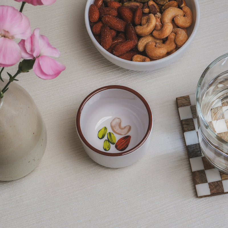 Looking down on the nuts bowl, showing a colorful illustration at the bottom of the bowl of pistachios, cashews, and almonds, and shown in view around the bowl are flowers, a larger bowl of nuts, and a glass of water.