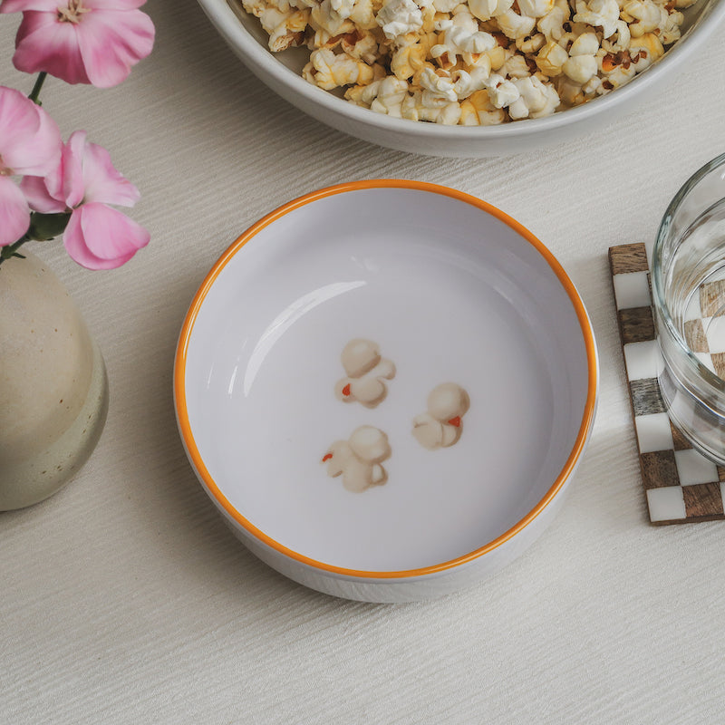 Looking down on the popcorn Macro Bowl showing a colorful illustration of popcorn on the bottom of the bow, with flowers, a larger bowl of popcorn, and a glass of water visible to the sides of the bowl.